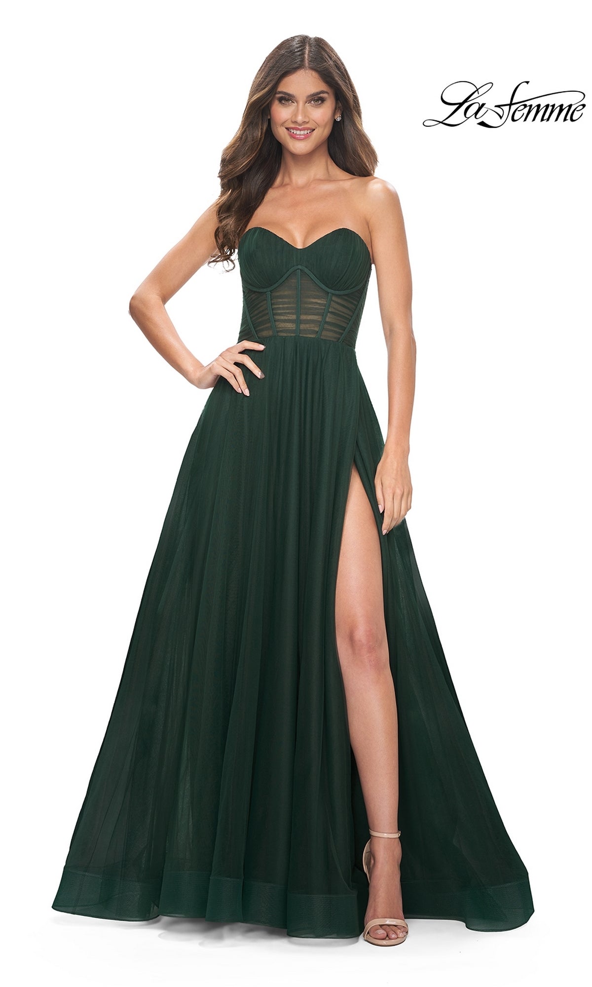 A woman stands smiling with one hand on her hip, wearing the La Femme Strapless Long A-Line Prom Dress 31971 in dark green, featuring a sheer corset bodice and a high leg slit.